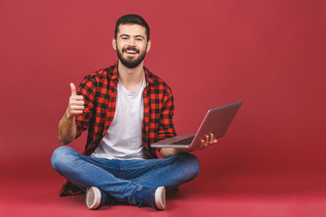 Portrait of a smiling young man in casual holding laptop computer while sitting on a floor and showing thumbs up gesture isolated over red background.