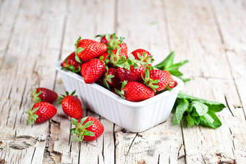 Fresh red strawberries in white bowl and mint leaves on rustic wooden background.