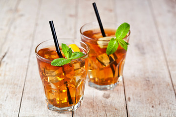 Traditional iced tea with lemon, mint leaves and ice cubes in two glasses on rustic wooden table.