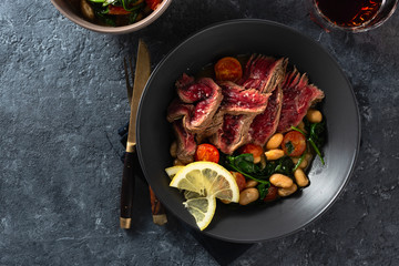 Dinner table. Beef steak with white beans, spinach and tomatoes on dark stone background with glass of red wine top view with free copy space