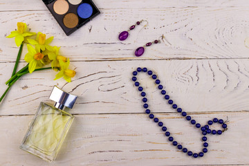 Women accessories on white wooden background. Bottle of perfume, eye shadow, necklace, earrings and yellow daffodils on white wood table. Beauty and fashion composition. Top view, flat lay