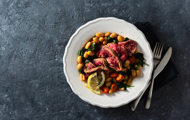 Dinner plate with beef steak with white beans, spinach and tomatoes on dark stone background top view with copy space