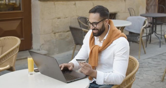 Handsome bearded eastern man makes online payment using credit card and laptop sitting outdoors cafe