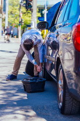 A sweaty tired young entrepreneur is wiping the sweat off his forehead while trying to change a flat tire on a beautiful sunny day, trouble in the city