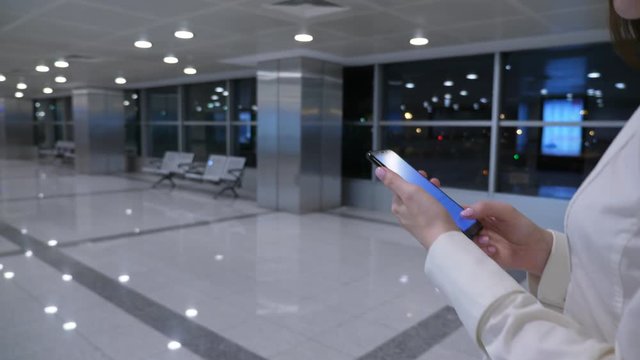 Woman type message using smartphone on the go, walking at airport terminal sterile area after arrival. She proceed to connecting flight, late evening time, empty passage