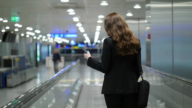 Business woman read news or checking email on the go using smartphone, terminal airside interior. Lady stay at moving travolator, go to boarding gate through empty airport, follow camera, blurred back
