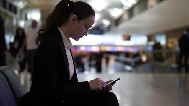Woman scroll through gallery on smartphone, blurred airport terminal hall on background, passengers pass by. Slow motion shot, dim interior at evening time. Lady spend time while waiting for flight