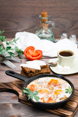 Ready-to-eat breakfast: shakshuka from fried eggs with tomatoes and parsley in a pan, bread with butter and coffeee on a wooden table
