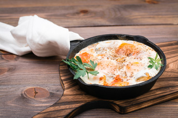 Homemade breakfast of shakshuka fried eggs with tomatoes and herbs in a pan on a wooden table