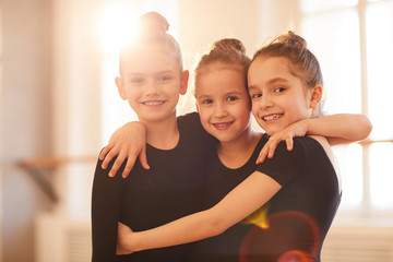 Portrait of three happy little girls embracing while posing in ballet studio lit by sunlight, copy...