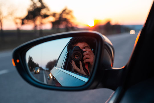 Young photographer taking selfie picture in a mirror of car with professional dslr camera, on background of sunset.