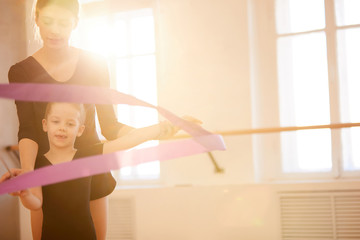 Portrait of coach teaching little girl doing gymnastics moves with ribbon in studio lit by warm...
