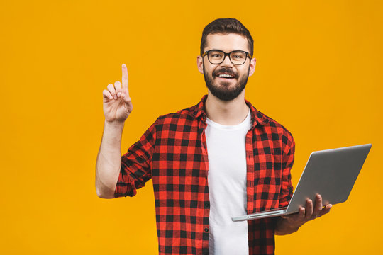 Young Bearded Man Holds Laptop And Finger Point Up Isolated Against Yellow Background.