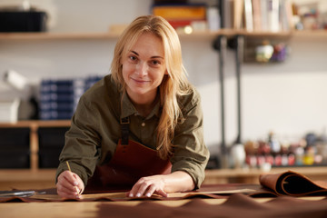 Portrait of female artisan smiling at camera while working with leather in workshop, copy space - obrazy, fototapety, plakaty