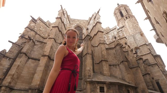 Tourist woman enjoy Barcelona Cathedral, stay against old building, turn head and point by hand, low angle portrait shot. Stone apse construction and bell tower seen on background