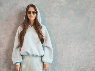 Young beautiful smiling woman looking at camera. Trendy girl in casual summer hoodie and skirt clothes. Funny and positive female posing near gray wall in studio. In sunglasses