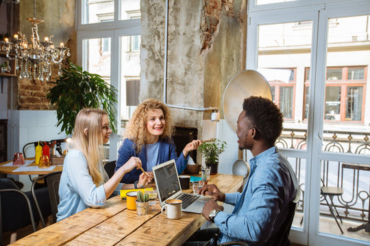 Two successful female friends meeting with african american man designer in modern cafe interior. Multiracial group of three friends having a coffee together. Concept with real people models.