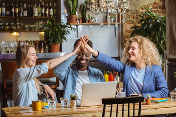 Celebrating success. Good job. African american man and two blond beautiful ladies giving high five. Couples sitting at the table with laptops and glasses of water in modern cafe interior. Copy space.
