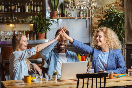Celebrating success. Good job. African american man and two blond beautiful ladies giving high five. Couples sitting at the table with laptops and glasses of water in modern cafe interior. Copy space.