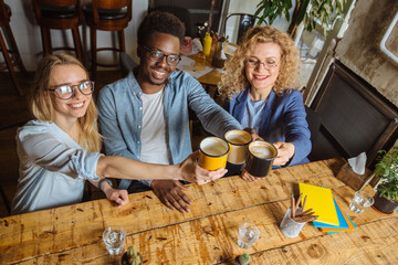 Top view of three multiethnic friends drinking latte at coffee bar restaurant. People in eyeglasses works, sits at wooden table clinking cups together at cafeteria. Friendship concept