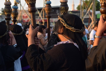 Unidentified Women penitents in garb and crown of bitter herbs carrying angel figurine scepter standing on the churchyard on Lenten procession.
