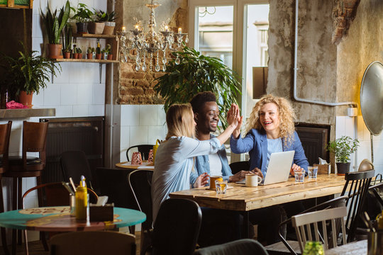 Celebrating success. Good job. African american man and two blond beautiful ladies giving high five. Couples sitting at the table with laptops and glasses of water in modern cafe interior. Copy space.