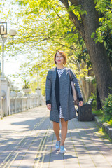 Fototapeta premium A beautiful woman with a short haircut in a dress walks down the street with a bag on her shoulder and looks thoughtfully into the distance with a smile.