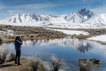 Fototapeta na wymiar Asian man tourist and photographer holding DSLR camera taking photo of Mammoth mountain landscape from the lake in Hot Creek Geological Site in winter. Travel photography concept
