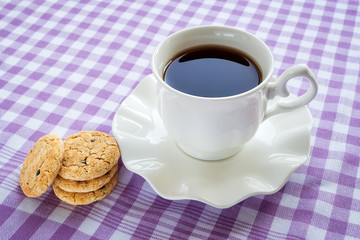 White porcelain saucer with wavy edge with cup of tea on it. Sweet oat cookies with pieces of dark chocolate. Breakfast on a checkered white purple tablecloth.