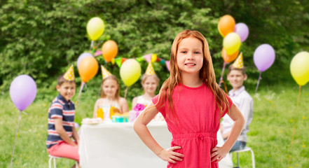 childhood and people concept - smiling red haired girl posing in pink dress over birthday party at summer park background