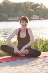 Fototapeta na wymiar Attractive smiling female sitting in lotus yoga position, wearing tight black clothes, hands on bare feet, stretching pose, outdoor, water in backdrop. Healthy lifestyle, keep fit, weight loss concept