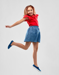 childhood, fun and motion concept - happy smiling girl in red shirt and skirt jumping over grey...
