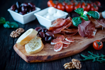 Fototapeta na wymiar Prosciutto, bread, olives, walnut, mozzarella, salami, basil and cherry tomatoes on brown wooden board. Mediterranean kitchen.