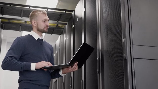 Server room in data center and support administrator typing at laptop. Male IT Technician engineer working with information standing before servers rows and checking the system using the notebook