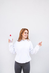 Fototapeta premium Happy, attractive girl on a white background, holding a bottle of water in her hands and listening to music in the headphones and looking away. Portrait of joyful fitness girl.
