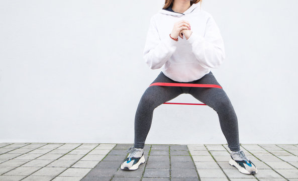 Cropped photo of fitness girl in stylish sportswear squats with a rubber band on the background of a white wall.Sporty athletic woman in sneakers and tracksuit squatting doing sit-ups in gym