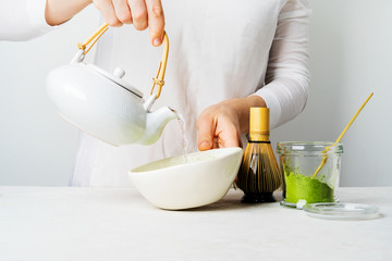 A woman in white pours water from a teapot to make organic Japanese green tea Matcha. Background with tools Chasen bamboo whisk, Chashaku spoon and bowl for brewing with copy space.