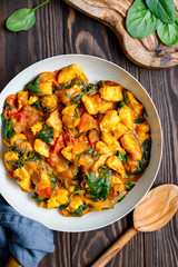 Top view on Bombay chicken curry with spinach, tomato and onion in a white frying pan on a kitchen wooden table. Indian traditional cuisine.