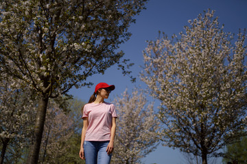Successful business woman enjoys her leisure free time in a park with blossoming sakura cherry trees wearing jeans, pink t-shirt and a fashion red cap