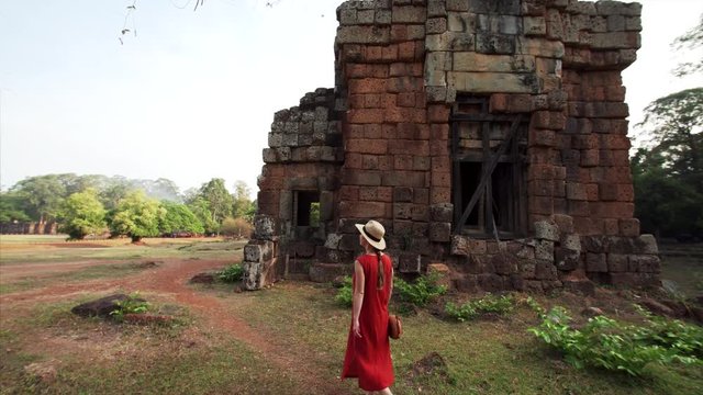 Woman in red dress and straw hat is walking near brick ruined Prasat Suor Prat, a series of twelve towers in Angkor Wat complex, made from rugged laterite and sandstone. Cambodia