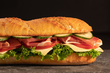 Huge fresh crispy baguette sandwich with meat, prosciutto, cheese, lettuce salad and vegetables. Close up. Black background. Space for text.