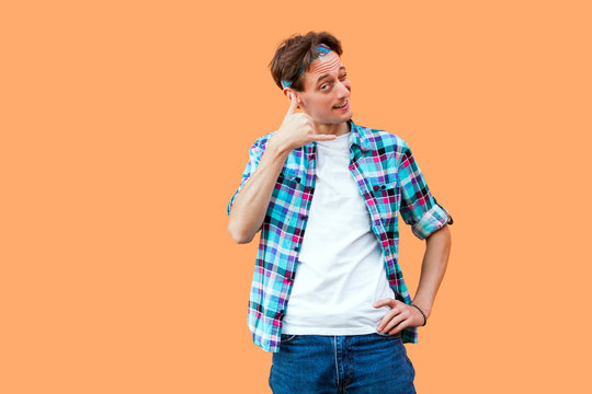 Portrait of funny young man in casual blue checkered shirt and headband standing with call gesture hand near ears and looking at camera winking. indoor studio shot, isolated on orange background.