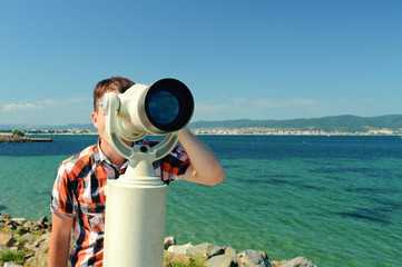 Obraz premium young man, traveler, looks through binoculars with coins against the backdrop of the landscape with the sea and mountains