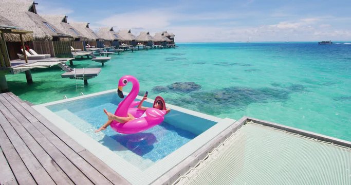 Vacation woman on inflatable Flamingo float mattress using mobile cell phone in swimming pool in overwater bungalow. Girl relaxing sunbathing enjoying travel at resort in bikini. Luxury lifestyle.