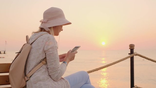 A stylish girl in pink clothes listening to music on a smartphone and sitting on a bench on a sea pier in the rays of a pink sunset