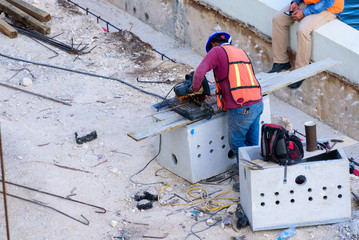 A latin worker cutting steel and another resting with his cell phone in hand in building construction, aerial view