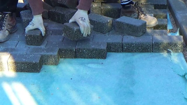Construction worker laying interlocking paving concrete onto sheet nonwoven bedding sand and fitting them into place.