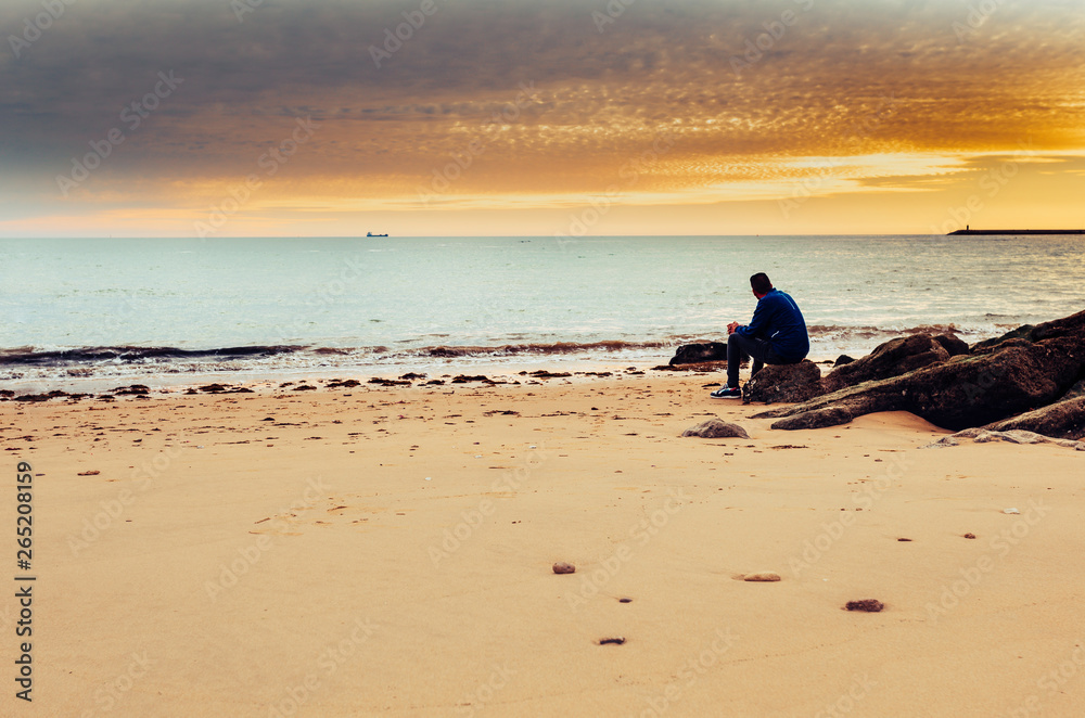 Wall mural Caucasian man alone sitting on the shore of the sea, contemplating the sunset