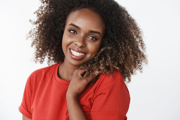 Close-up shot of carefree happy and gentle gorgeous african-american female model with curls smiling broadly touching hair, gazing delighted, tender at camera wearing red t-shirt over white background