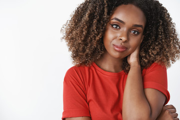 Close-up shot of tender and gentle romantic african-american girlfriend in red casual t-shirt touching back of neck shy and cute tilting head, smiling sensually with flirty gaze over white background